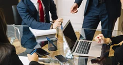 Vier Personen in Businesskleidung sitzen an einem Glastisch mit Laptop, Smartphone, Notizblock und Stiften, eine Person steht und gestikuliert.