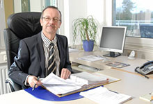 Jürgen Reinhardt ist Ansprechpartner in Erlangen und sitzt in Anzug und Krawatte an Schreibtisch mit Aktenordnern, Computerbildschirm und Telefon, Büro mit Fenster und Pflanze im Hintergrund.